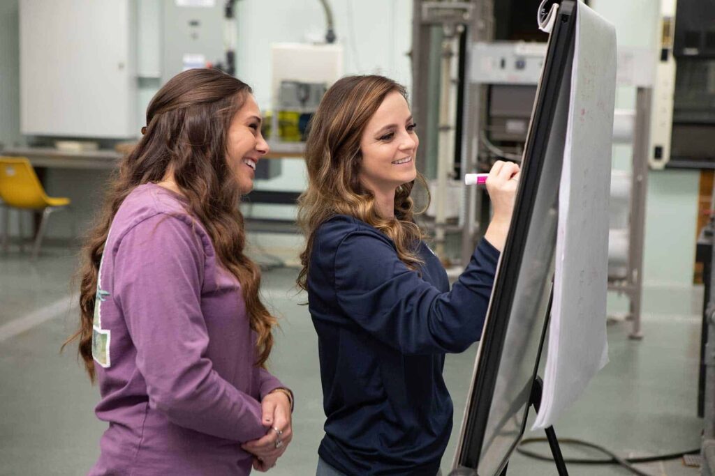 Two women collaborating in a workshop, writing on a flipchart, smiling and engaged in discussion.