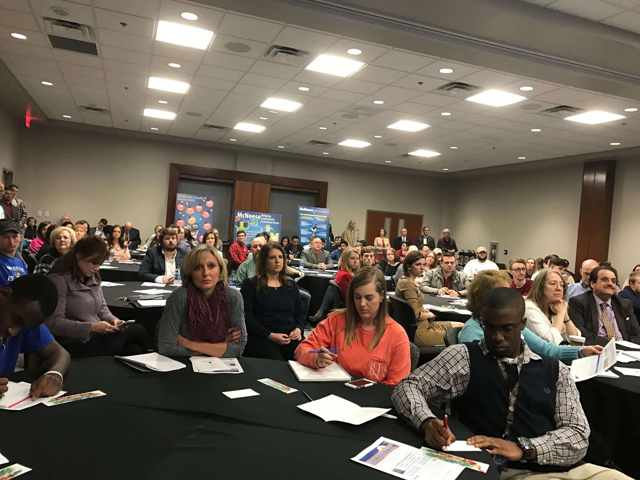 Audience attentively listens at a conference in a large room with tables, papers, and promotional banners in the background.