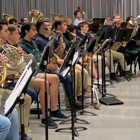 Students performing in a school band with various brass and wind instruments in a rehearsal room.