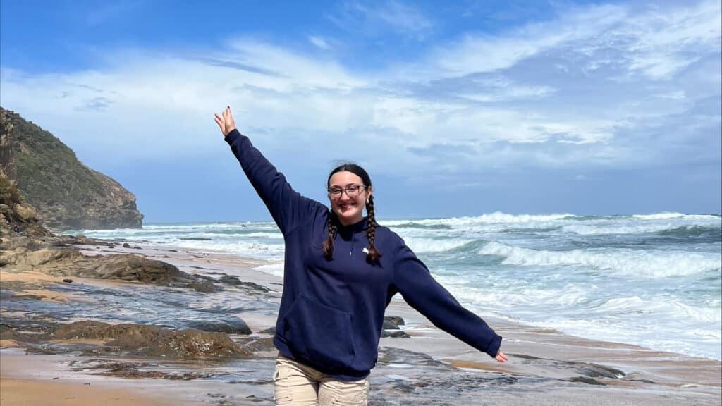 Young woman with braided hair poses with her hands up and a big smile on a beach with waves and mountains in the background.
