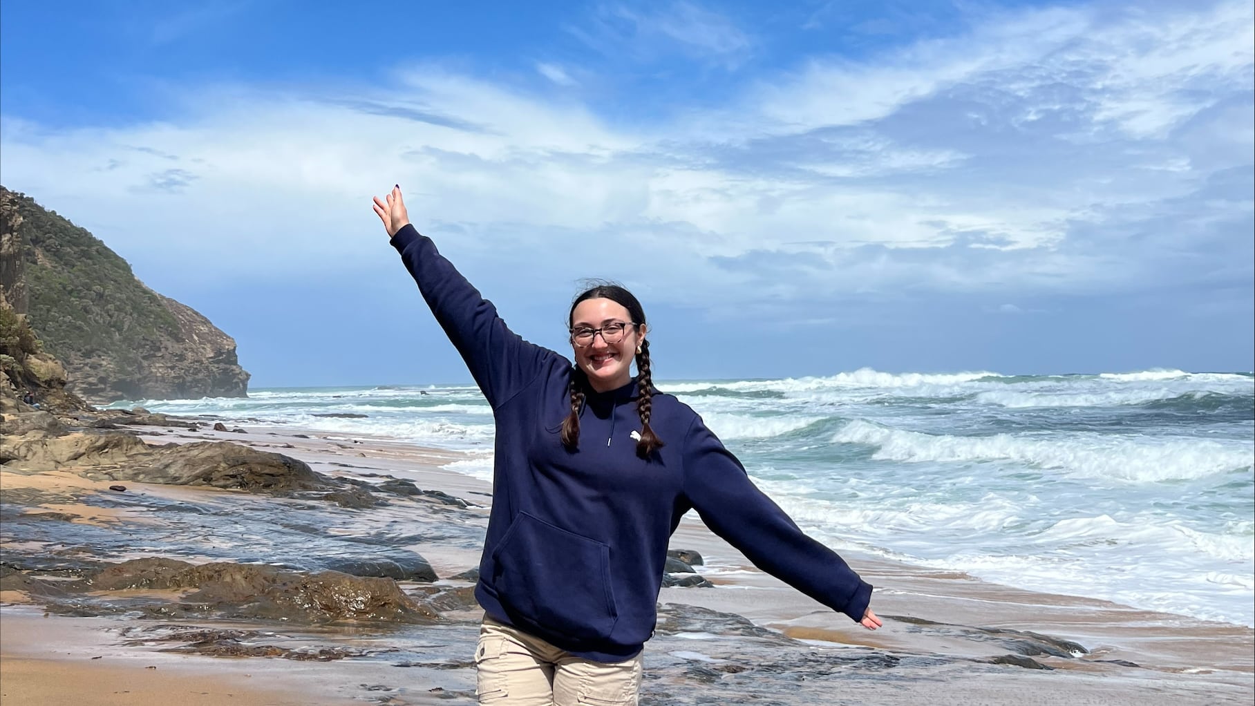 Young woman with braided hair poses with her hands up and a big smile on a beach with waves and mountains in the background.
