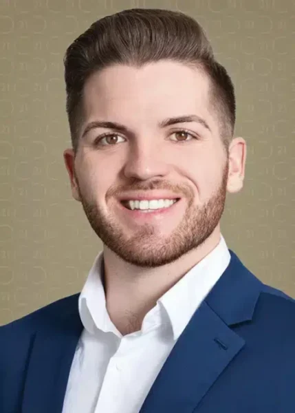 Smiling man with brown hair and beard wearing a blue suit and white shirt, against a beige background.