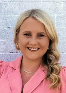 Smiling person in a pink blouse against a light brick wall, blonde hair styled in loose waves.