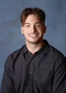 Smiling man with short hair and a black button-up shirt against a blue background.