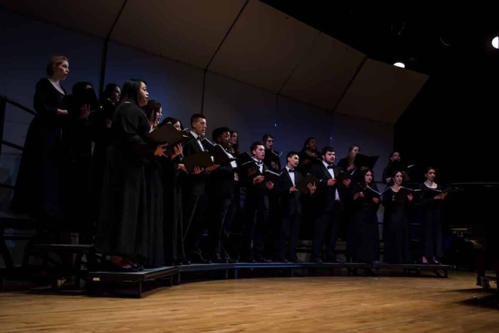 Choir dressed in formal black attire performs on stage in dim lighting.
