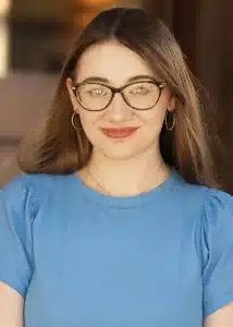 Woman with glasses and long hair wearing a blue shirt, smiling confidently.