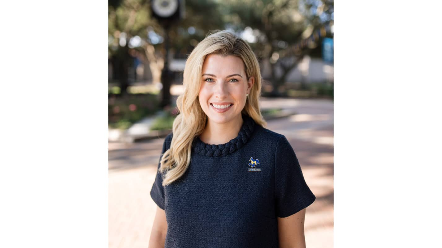 Woman smiling outdoors wearing navy dress with embroidered logo, trees in background.