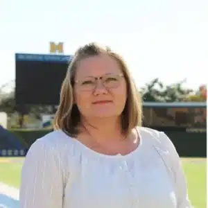Woman in white blouse at university sports field with scoreboard in background.