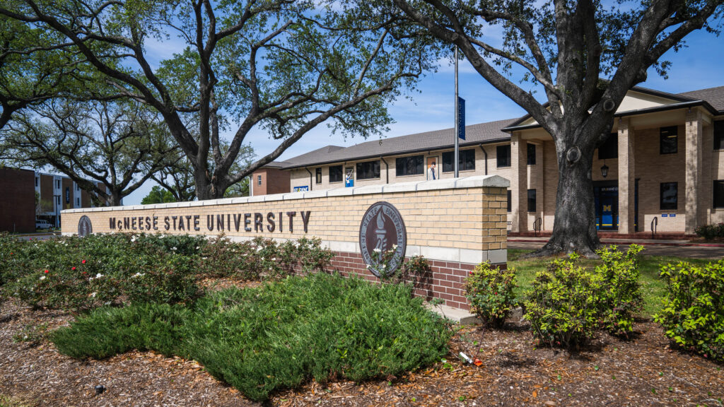 McNeese State University entrance sign surrounded by greenery and large oak trees under a clear blue sky.