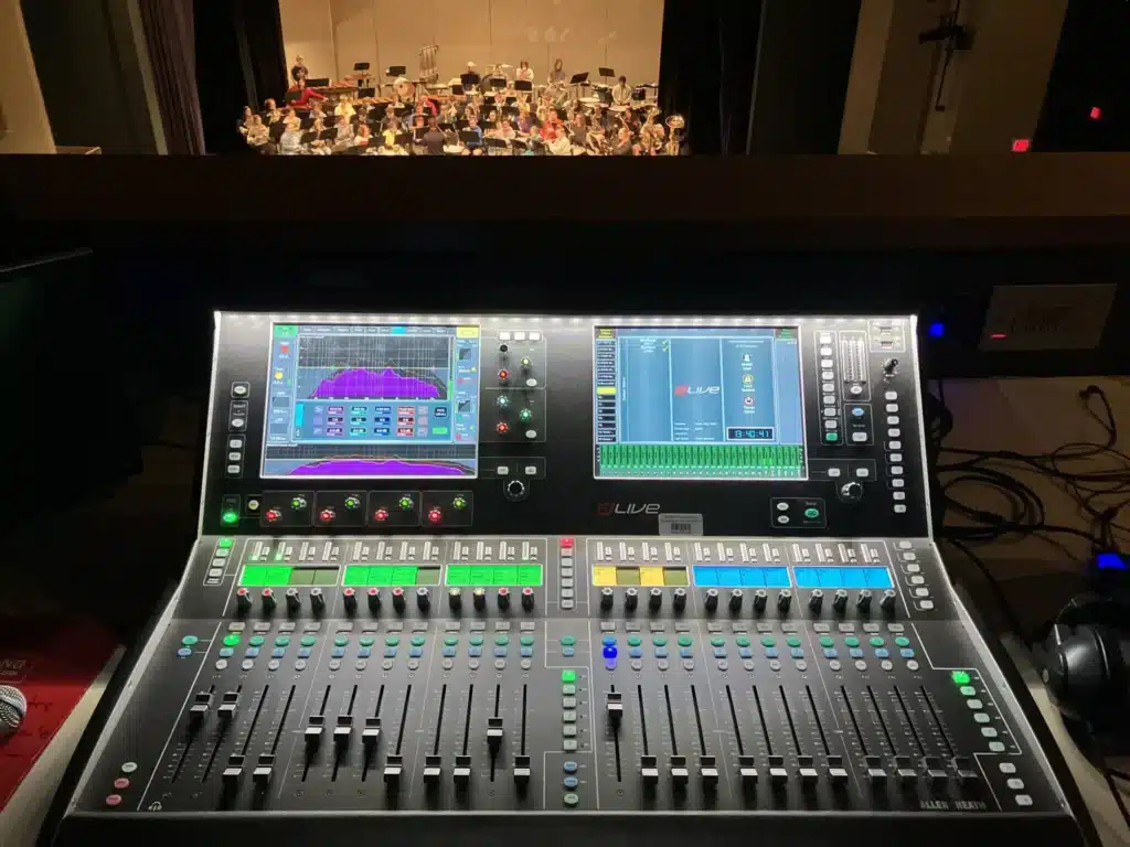 closeup of sound mixing board with video displays in a sound booth overlooking a theatre stage
