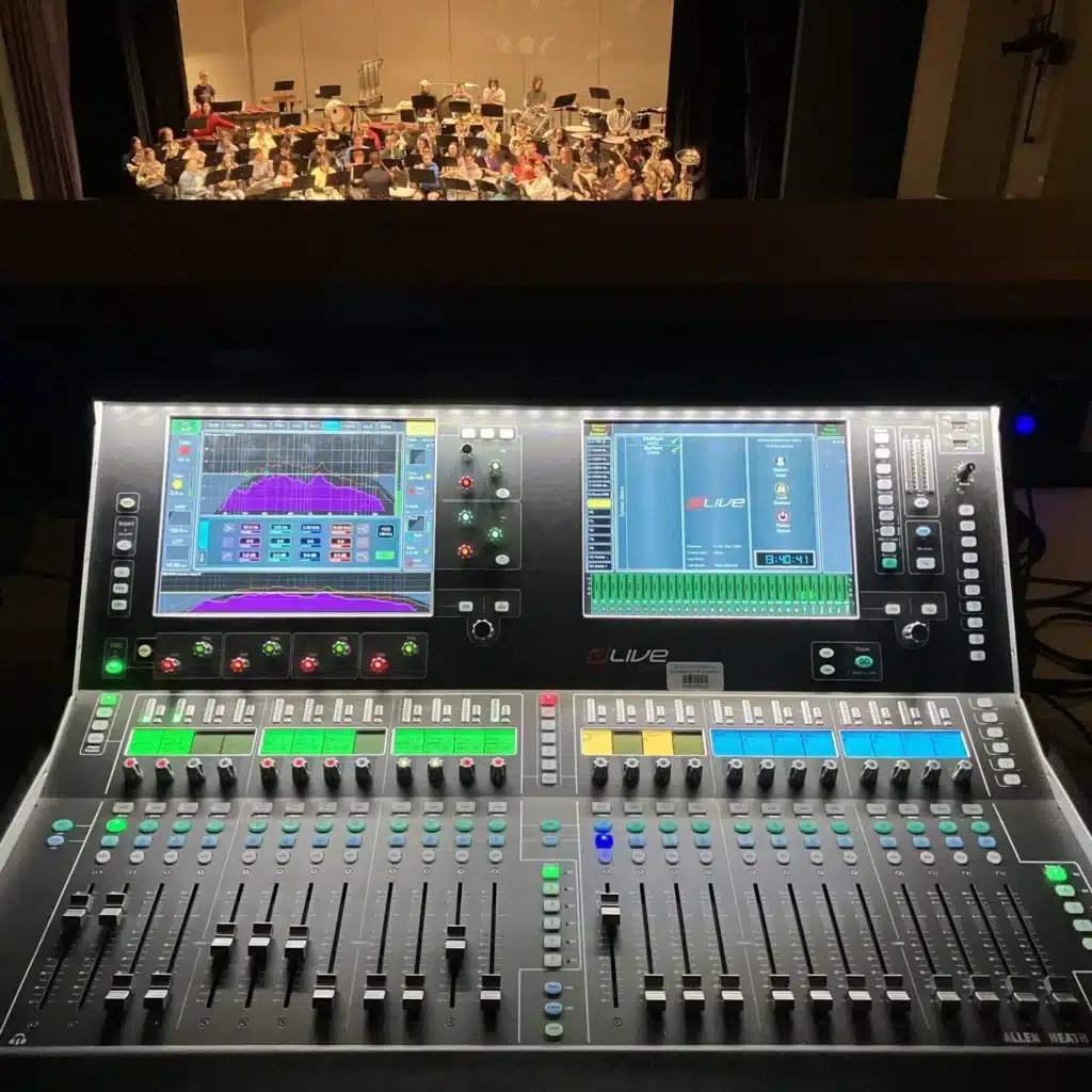 Digital mixing console in a theater with an orchestra performing in the background.