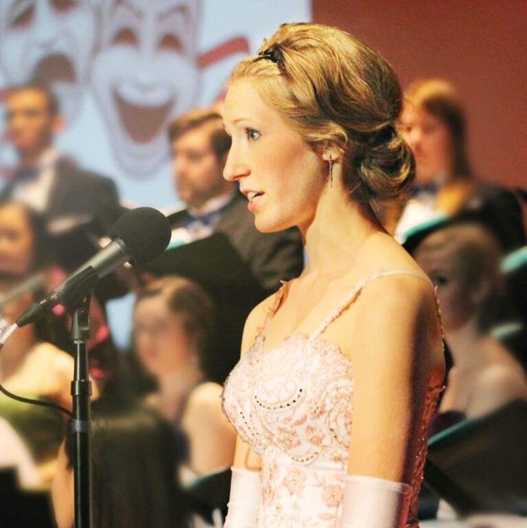 Woman in formal gown singing at a microphone in front of a choir