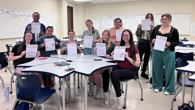 Group of students in a classroom holding signs with the word 'READ', smiling and posing around a table.