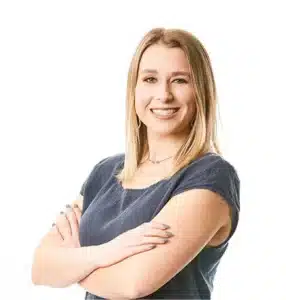 Smiling woman with blonde hair crosses arms, wearing a blue top against white background.