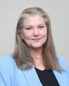 Smiling woman with long hair in a light blue blazer and black top against a neutral background.