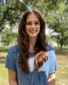 Smiling woman with long brown hair in blue blouse outdoors with trees in background.