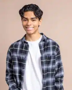 Smiling person wearing a plaid shirt and white t-shirt against a neutral background.