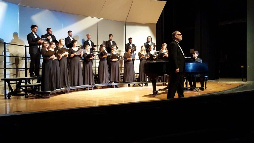 Choir performing on stage with conductor and pianist, choir members in black attire holding sheet music.