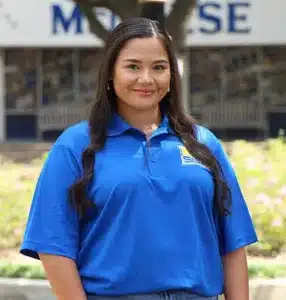 Smiling woman in blue polo shirt standing outdoors with blurry building in background.