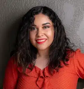 Woman with long curly hair wearing a red blouse and smiling against a textured gray wall.