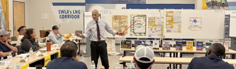 Norman Lieberman presents a troubleshooting process operations seminar at the McNeese State University LNG Center of Excellence while standing in front of illustrated process diagrams. Participants seated around classroom tables listen attentively and take notes during the three-day industry training.