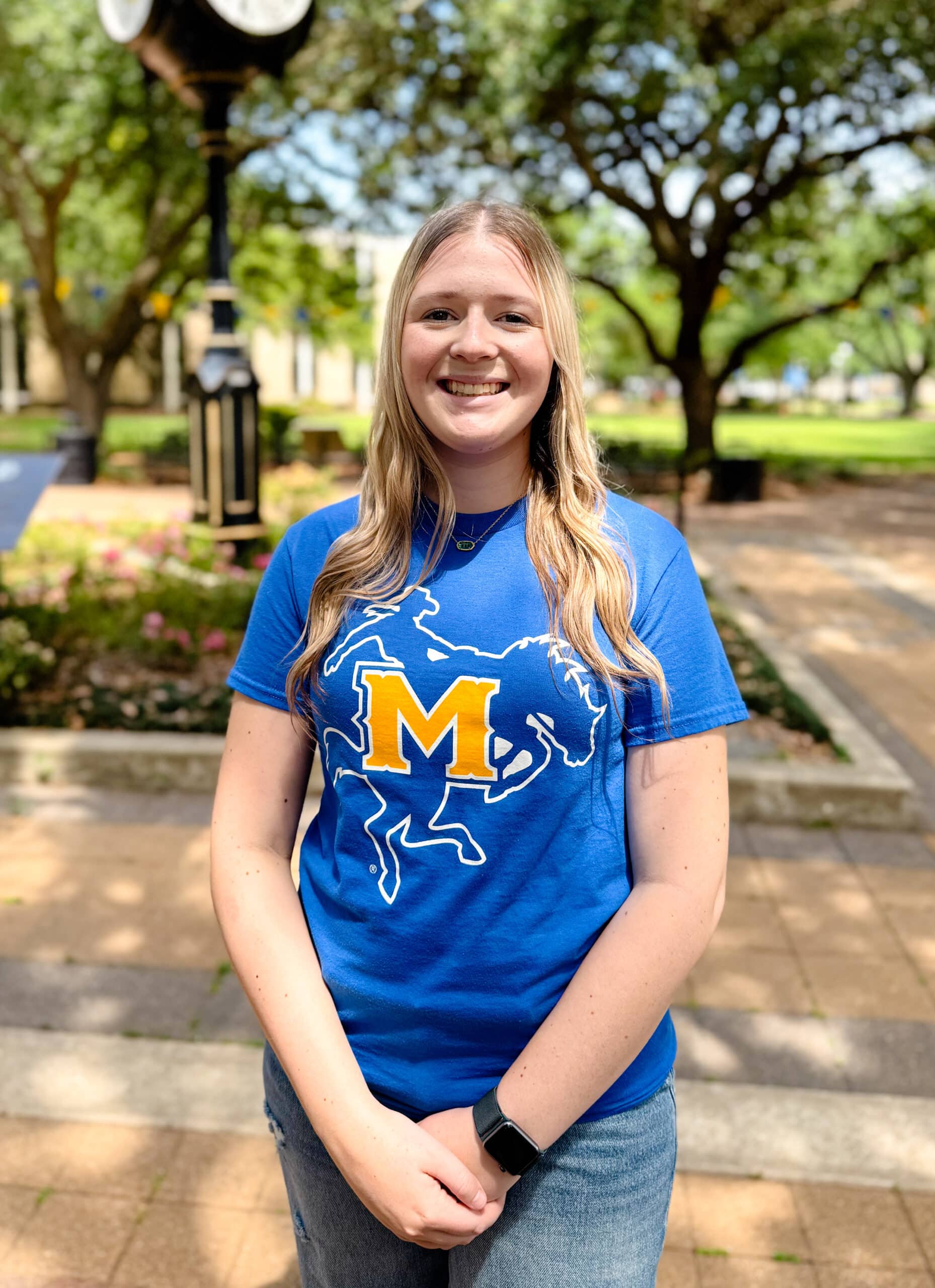 Lylie Loftis smiling outdoors in front of Clock Tower, wearing a blue and yellow T-shirt.